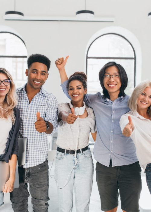 Blonde female student in ripped jeans holding laptop and laughing after hard test. Asian guy in glasses waving hands, celebrating end of exams and beginning of vacation.
