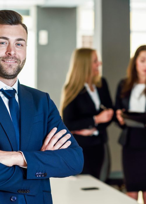 Caucasian businessman leader looking at camera in modern office with multi-ethnic businesspeople working at the background. Teamwork concept. Young man with beard wearing blue suit.