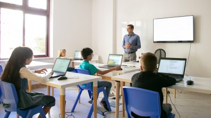 Confident teacher explaining lesson to pupils. Multiethnic children sitting at table in classroom, listening middle-aged man and using laptop computers. Childhood and digital education concept